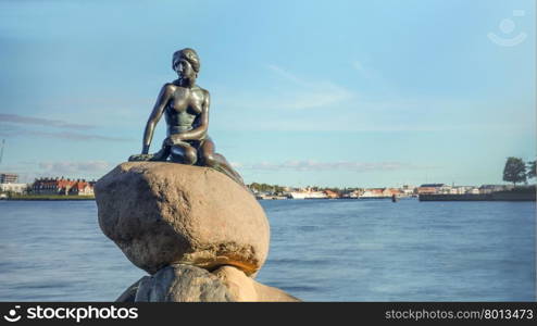 Front view of Little Mermaid statue on large boulders in Denmark with harbor under blue sky in the background. Little Mermaid statue on rock in Denmark