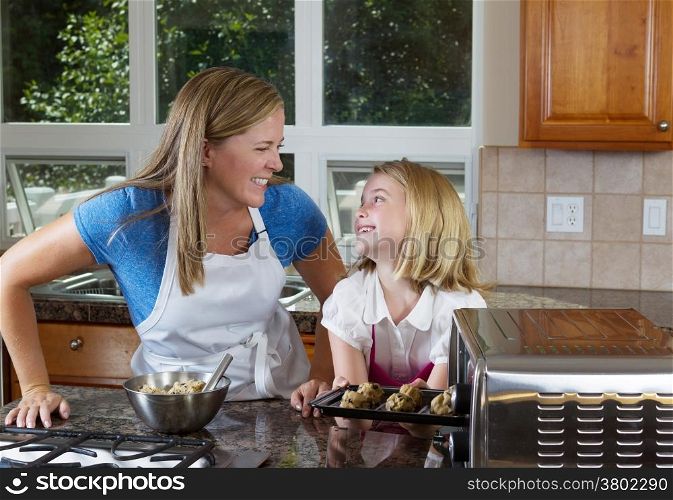 Front view of mother and her young daughter looking at each other as they put raw cookie dough into portable oven