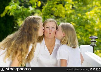 Front view of mother, displaying fish lips, being kissed by her two daughters outdoors on patio with woods in background
