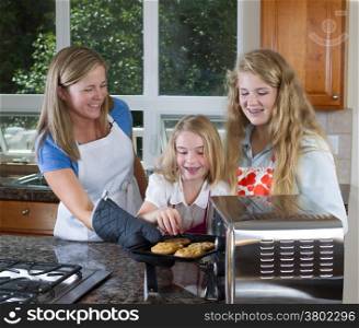 Front view of mother pulling out fresh cookies from the oven with her youngest daughter reaching for them
