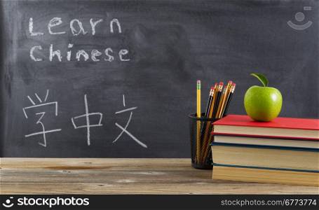 Front view of rustic desk for student learning Chinese with books, pencils and a green apple in front of chalkboard with Mandarin text.