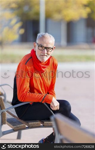 Front view of senior man in sports clothes sitting in a bench at park while smiling and looking camera in a sunny day