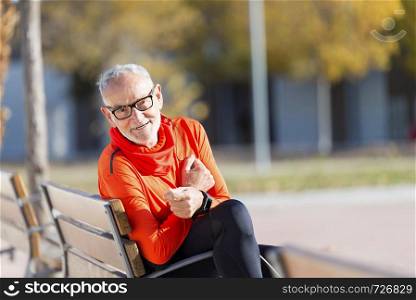 Front view of senior man in sports clothes sitting in a bench at park while smiling and looking camera in a sunny day