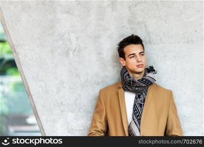 Front view of stylish young man wearing coat and scarf leaning on a wall while looking away
