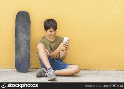 Front view of young boy sitting on ground leaning on a yellow wall while using a mobile phone