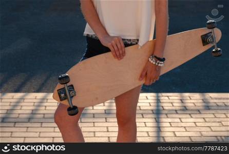 Front view of young girl in shorts holding longboard by both hands. Skateboard Extreme Sport Skater Activity Concept. Shadow pattern on pavement behind model.. Skateboard Extreme Sport Skater Activity Concept. Front view of young girl in shorts holding skateboard.