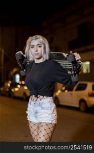 Front view of young pretty woman looking camera in shorts while standing on the street holding a skateboard at night in the city
