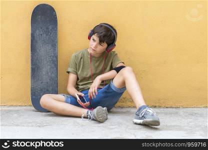 Front view of young skater boy sitting against a yellow wall while listening music by headphones