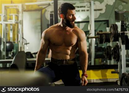 Front view portrait of adult caucasian man male athlete muscular bodybuilder or fitness trainer with black hair and beard sitting shirtless at the gym looking to the side resting