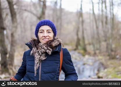 Front view portrait of beautiful girl with backpack standing near old footbridge. She is having break during hike in coniferous forest