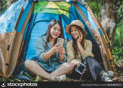 front view Young Asian pretty woman and her girlfriend sitting at front of tent, use mobile phone take photo during c&ing in forest with happiness together