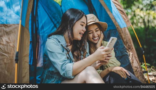 front view Young Asian pretty woman and her girlfriend sitting at front of tent, use mobile phone take photo during c&ing in forest with happiness together