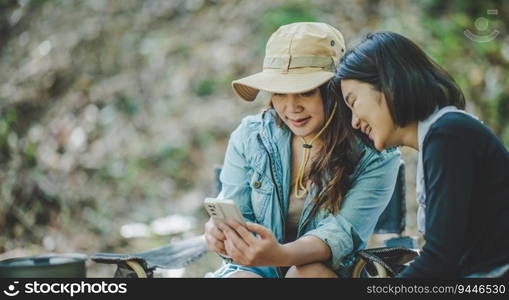 front view Young Asian pretty woman and her girlfriend sitting at front of tent, use mobile phone take photo during c&ing in forest with happiness together