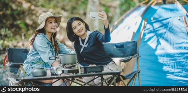 front view Young Asian pretty woman and her girlfriend sitting at front of tent, use mobile phone take photo during c&ing in forest with happiness together