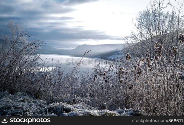 Frosted winter landscape in cold temperature with ice and hoar frost on lake, rime on crisp, frozen leaves and mountains in the horizon at sunset with sun rays.