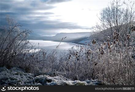 Frosted winter landscape in cold temperature with ice and hoar frost on lake, rime on crisp, frozen leaves and mountains in the horizon at sunset with sun rays.