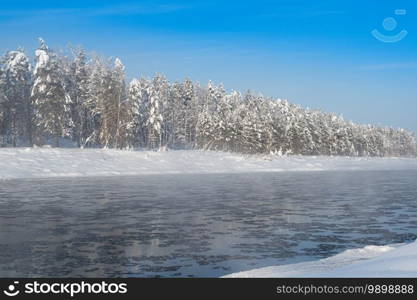 Frosty fog over winter river with snow and forest on bank. First ice on lake on cold day.