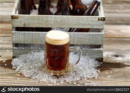Frosty glass mug of dark beer, focus on front, with vintage crate filled with bottled beer and crushed ice on rustic wooden boards.
