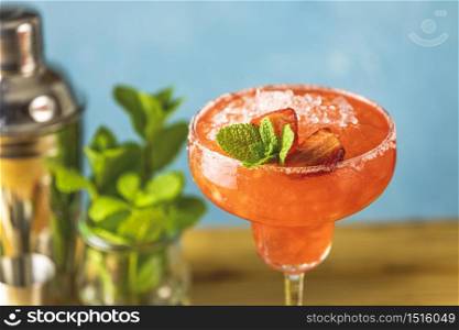 Frozen strawberry lime mint margarita in tall footed glass close up on the wooden table with bar tools and ingredients. Luxury alcohol fresh drink.