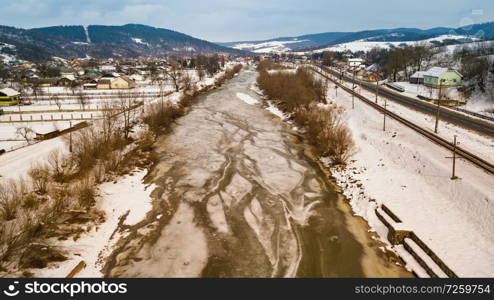 Frozen winter river. Aerial rural view of snowy village, road and railway. Melting snow and ice in early spring.