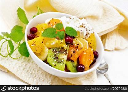 Fruit salad with banana, orange, kiwi, cranberries and baked pumpkin, whipped cream, sprinkled with chocolate and coconut with mint in a bowl on a napkin on wooden board background