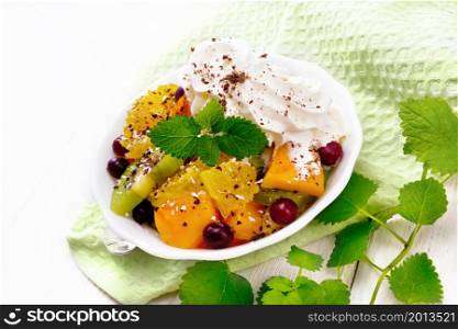 Fruit salad with orange, kiwi, cranberries and baked pumpkin, whipped cream, sprinkled with chocolate and coconut with mint in a bowl on a napkin on white wooden board background