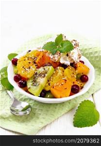 Fruit salad with orange, kiwi, cranberries and baked pumpkin, whipped cream, sprinkled with chocolate and coconut with mint in a bowl on a napkin on the background of light wooden board