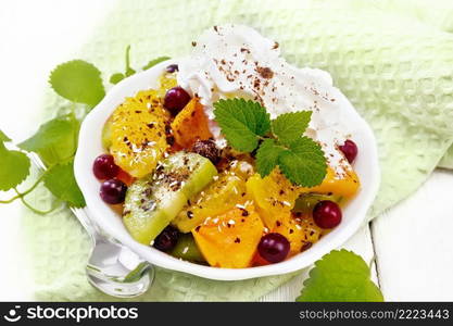 Fruit salad with orange, kiwi, cranberries and baked pumpkin, whipped cream, sprinkled with chocolate and coconut with mint in a bowl on a towel against the background of light wooden board