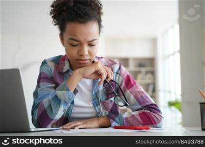 Frustrated afro girl is overworked studying remote at home. Teenage hispanic schoolgirl is sitting at the desk in front of laptop tired from homework. Stressed student has problems with exam.. Frustrated afro girl is overworked studying remote at home. Schoolgirl is tired from homework.