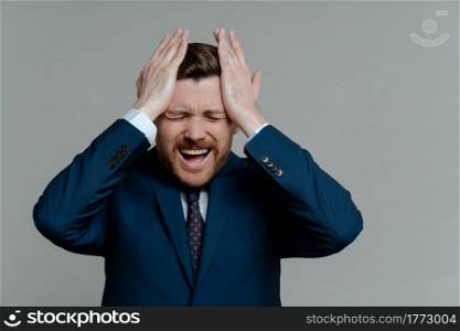 Frustrated business man in suit feeling stressed and worried of financial problems, guy touching head with hands while getting bad news while standing against grey background. Business failure concept. Upset businessman feeling stressed of failure at work