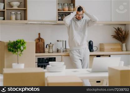 Frustrated Caucasian man faces financial problems, rents new apartment, looks with puzzlement at laptop computer, dressed in casual wear, keeps hands on head, poses against kitchen interior.