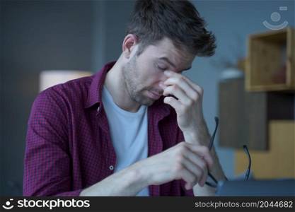 Frustrated male Austrian entrepreneur tired of remote work online sitting at modern home office, overworked man removing glasses massages bridge of his nose suffering from eye pressure and fatigue. Frustrated male Austrian entrepreneur tired of remote work online sitting at modern home office