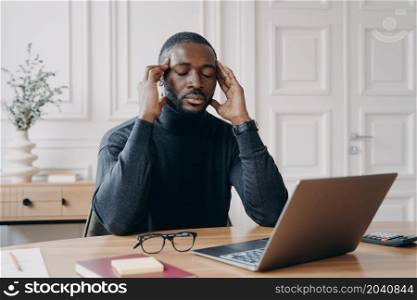 Frustrated tired afro american man office worker with closed eyes trying to concentrate while sitting at workplace, african ethnicity male employee massaging temples, suffering from headache at work. Frustrated tired afro american man office worker with closed eyes trying to concentrate at workplace