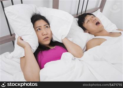 Frustrated woman covering ears with pillow while man snoring in bed