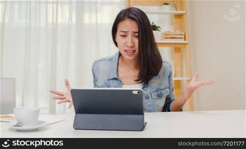 Frustrated Young Asia lady having problem with not working tablet computer sitting on desk. Freelance smart business women casual wear using tablet working in workplace in living room at home office.