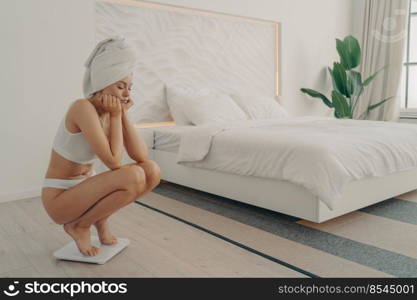 Frustrated young caucasian girl standing barefoot squatted on electronic scale in underwear after taking shower in modern bedroom next to large bed, feeling sad. Weight loss and dieting concept. Frustrated young caucasian girl standing barefoot squatted on electronic scale