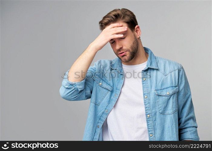 Frustrated young caucasian man in jeans shirt touching head with hand while standing overg studio grey background.. Frustrated young caucasian man in jeans shirt touching head with hand while standing overg studio grey background