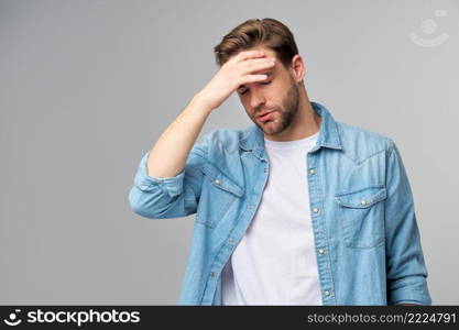 Frustrated young caucasian man in jeans shirt touching head with hand while standing overg studio grey background.. Frustrated young caucasian man in jeans shirt touching head with hand while standing overg studio grey background