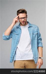 Frustrated young caucasian man in jeans shirt touching head with hand while standing overg studio grey background.. Frustrated young caucasian man in jeans shirt touching head with hand while standing overg studio grey background