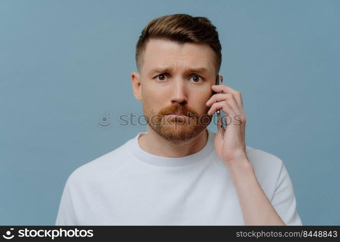 Frustrated young man in white shirt receiving bad unpleasant news on mobile phone and feeling upset, looking at camera with unhappy facial expression while standing against blue studio background. Worried man receiving bad news while talking with somebody on mobile phone