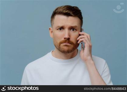 Frustrated young man in white shirt receiving bad unpleasant news on mobile phone and feeling upset, looking at camera with unhappy facial expression while standing against blue studio background. Worried man receiving bad news while talking with somebody on mobile phone