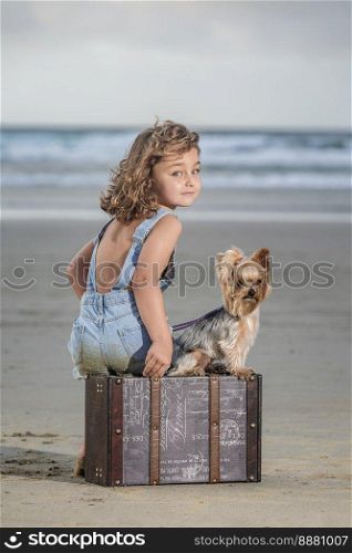 Full body adorable girl and Yorkshire Terrier dog sitting on suitcase and looking at camera while spending stormy weekend day on Famara Beach in summer in Lanzarote, Spain. Girl with dog sitting on luggage near sea