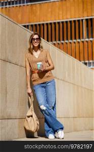 Full body adult woman in stylish clothes with bag and coffee to go leaning on wall and looking away on city street. Stylish female with coffee leaning on wall