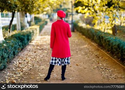 Full body back view of anonymous female in red outfit standing on pathway in park with trees on autumn day. Unrecognizable stylish woman in an urban park
