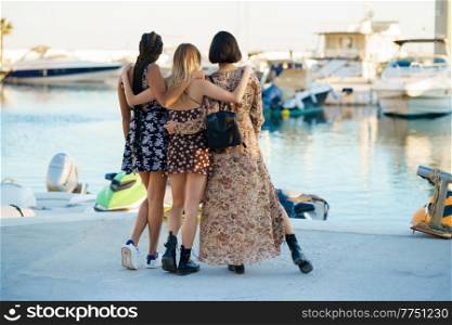 Full body back view of multiethnic unrecognizable female friends embracing gently while standing on seafront and observing marina. Stylish diverse women hugging on pier with moored boats