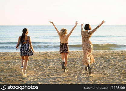 Full body back view of unrecognizable female friends in dresses running on sandy beach with raised arms near waving sea. Faceless girlfriends walking towards sea