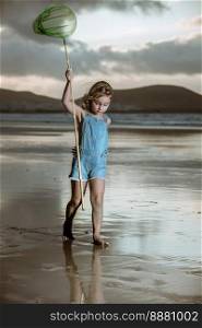 Full body barefoot girl in denim overall shorts raising arm with scoop net walking on wet sand near waving sea at sunset on Farmara Beach in Lanzarote, Spain. Girl with scoop net walking near sea