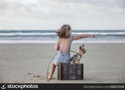 Full body barefoot girl in denim overall shorts sitting on suitcase and pointing away for attentive Yorkshire Terrier dog while spending stormy day on sandy Famara Beach in Lanzarote, Spain. Girl pointing away for adorable dog