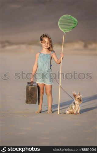 Full body barefoot girl in denim overall shorts with luggage and scoop net standing on sand near Yorkshire Terrier dog on leash on summer day Famara Beach in Lanzarote, Spain. Girl with suitcase and scoop net standing near dog on beach