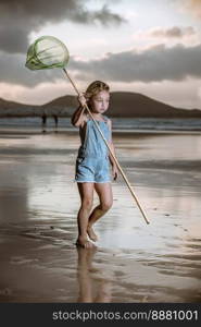 Full body barefoot girl in denim overall shorts with scoop net on long handle looking away and walking on wet sand against sea and cloudy sundown sky in weekend on Famara Beach in Lanzarote, Spain. Girl with scoop net exploring beach at sunset